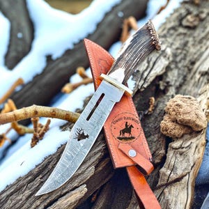 May include: A handcrafted hunting knife with a patterned blade and antler handle rests in a brown leather sheath. The blade features a deer silhouette and the text "Cowboy BladeWorks" is visible on the sheath.