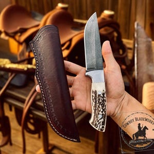 May include: A handcrafted hunting knife with a textured blade and a bone handle. The knife is held next to a dark brown leather sheath with decorative stitching. The image also includes the "Cowboy Bladeworks" logo.