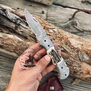 May include: A folding knife with a patterned steel blade and a handle featuring a combination of patterned metal and a tan and brown antler design. The knife is held in a hand, with the "Cowboy Bladeworks" logo visible.