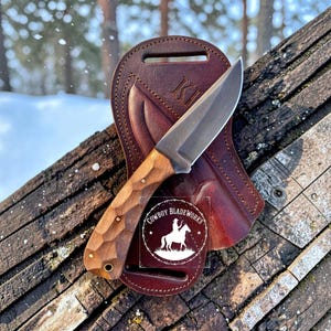 May include: A fixed-blade knife with a textured wooden handle and a silver metal blade, resting on a dark brown leather sheath. The sheath features a stamped logo with the text COWBOY BLADEWORKS and an illustration of a person on horseback.
