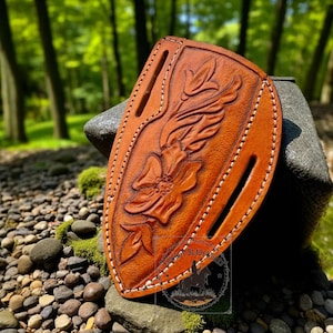 May include: A brown leather knife sheath with a floral design. The sheath has a decorative embossed flower and leaf pattern. White stitching outlines the edges and the sheath has two slots for a belt.