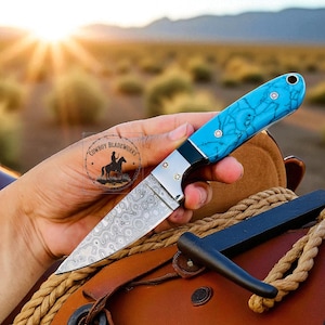 May include: A handcrafted knife with a patterned blade and a turquoise handle. The knife features a silver bolster and decorative rivets. The blade is held in a hand, with a blurred desert landscape in the background. The knife is resting on a brown leather saddle.