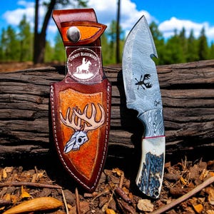 Puede incluir: Un cuchillo de caza artesanal con una hoja estampada y un mango de hueso, junto a una funda de cuero marrón. La funda presenta un diseño de calavera de ciervo y el texto "Cowboy BladeWorks". El cuchillo tiene un gráfico de ciervo.