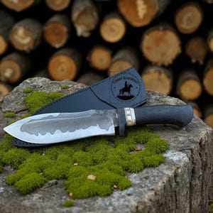May include: A handcrafted knife with a dark, textured handle and a silver blade, resting on a mossy surface. The knife is accompanied by a black leather sheath. The blade has a unique design, and the handle has a silver accent.