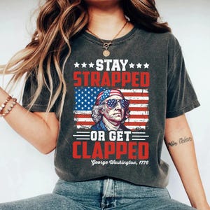 Könnte beinhalten: Dunkelgraues T-Shirt mit einer Grafik von George Washington mit Sonnenbrille vor einer amerikanischen Flagge. Der Text lautet "Stay Strapped or Get Clapped" und "George Washington, 1776".