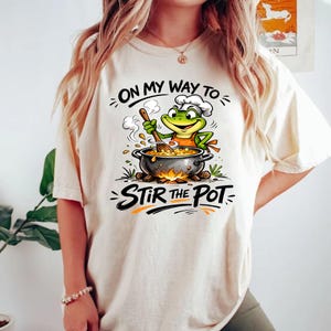 Puede incluir: Camiseta color crema con un dibujo animado de una rana chef removiendo una olla sobre un fuego. El texto "ON MY WAY TO" y "STIR THE POT" están encima y debajo de la ilustración de la rana. La rana lleva un gorro y un delantal de chef.