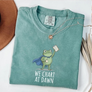 Op de afbeelding: Een teal Comfort Colors t-shirt met een cartoon kikker met een cape en een klembord en een koffiekopje. De tekst "WE CHART AT DAWN" staat eronder. Een zilveren ketting met een hanger ligt over het shirt.