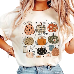May include: A cream-colored t-shirt featuring a graphic print of various pumpkins decorated with medical themes, including nurse caps, stethoscopes, syringes, bandages, a clipboard labeled nurse, and scrubs, interspersed with small stars and hearts.