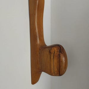 May include: A wooden wall hook with a natural finish. The hook has a curved, organic shape, featuring a rounded knob and a long, slender stem. The wood grain is visible, adding to its rustic charm.