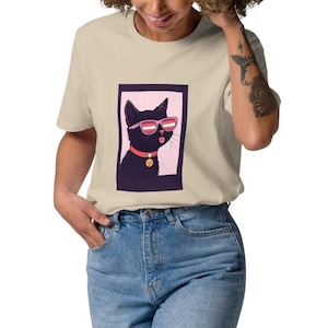 Puede incluir: Camiseta beige con un gráfico de un gato negro con gafas de sol con lentes rosas y un collar rojo con un dije dorado. El gato está dentro de un marco rectangular rosa y morado. La camiseta es de material suave.