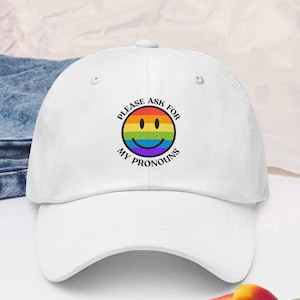 Könnte beinhalten: Weiße Baseballkappe mit gebogenem Schirm. Die Kappe hat ein kreisförmiges Design mit einem Regenbogen-Smiley und dem Text "PLEASE ASK FOR MY PRONOUNS" in Schwarz. Die Kappe ist aus einem weichen Material.