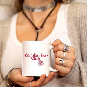 Pode incluir: Caneca de cerâmica branca com o texto "Carabin her Club" em escrita marrom. A caneca apresenta um gráfico de uma mão com os dedos apontando para cima. A caneca é segurada por uma pessoa usando vários anéis.
