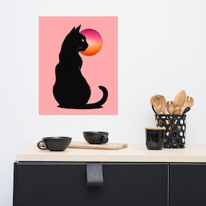 May include: A black cat silhouette art print against a pink background with a gradient orange and purple sun. The cat is sitting and looking to the left. The print is displayed above a black cabinet with black cups and wooden utensils.
