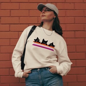 Pode incluir: Camisola de manga comprida creme com um gráfico de três gatos pretos espreitando por cima de um design de bandeira do orgulho lésbico. A camisola é feita de um material macio e tem um ajuste descontraído.