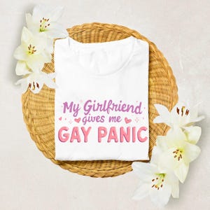May include: A white t-shirt with the text "My Girlfriend gives me GAY PANIC" in pink and purple lettering. The shirt is displayed on a woven basket, decorated with white flowers.
