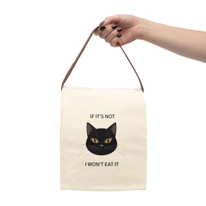 May include: A canvas lunch bag with a brown handle. The bag features a black cat face with yellow eyes and the text "IF IT'S NOT I WON'T EAT IT" in black font. The bag is held by a person.