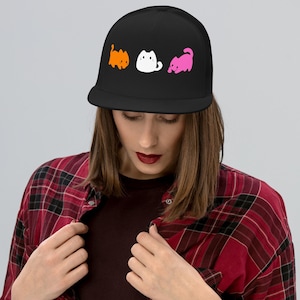 May include: Black flat-peak cap with three cartoon cats in orange, white, and pink. The cats are in different poses. The cap has a mesh back and a structured front. A stylish accessory for cat lovers.