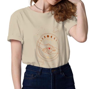 以下が含まれることがあります： ベージュの半袖Tシャツ。天体のデザインが特徴です。グラフィックには、月の満ち欠け、惑星、星に囲まれた、心臓を抱きしめる手が含まれています。柔らかい素材で作られたシャツです。