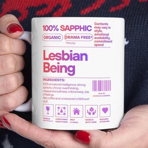 May include: White ceramic mug with the text "100% Sapphic" and "Lesbian Being" in purple and red. The mug includes a list of "ingredients" and icons. The mug is held by a person.
