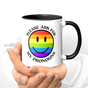 Puede incluir: Una taza de cerámica blanca con interior y asa negros. La taza presenta una carita sonriente con los colores del arcoíris y el texto "PLEASE ASK FOR MY PRONOUNS" en negro, promoviendo la inclusión y el respeto por la identidad de género.