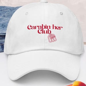 Pode incluir: Boné de beisebol branco com o texto vermelho "Carabin her Club". O design inclui um gráfico vermelho de uma mão segurando mosquetões. O boné é feito de um tecido macio e tem uma aba curva.