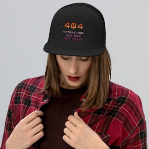 May include: Black trucker hat with the text "404 Attraction For Men Not Found" in orange and purple. The hat has a mesh back and a flat brim. The person in the image is wearing a red and black plaid shirt.
