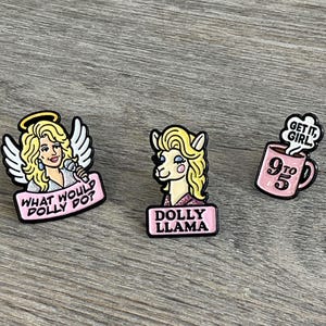 May include: Three enamel pins featuring Dolly Parton-inspired designs. One pin depicts Dolly with angel wings and the text "What Would Dolly Do?". Another shows a llama with Dolly's hair and the text "Dolly Llama". The third pin is a pink mug with "9 to 5" and "Get it, Girl!"