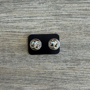 May include: Two silver-colored pin backs are displayed on a small black rectangular card. The pin backs have a butterfly clutch design. The background is a light gray wooden surface.