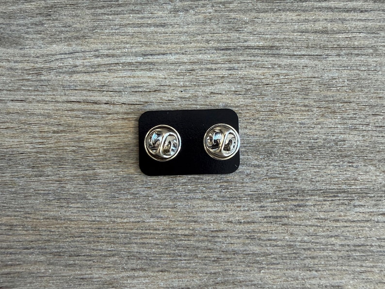 May include: Two silver-colored pin backs are displayed on a small black rectangular card. The pin backs have a butterfly clutch design. The background is a light gray wooden surface.