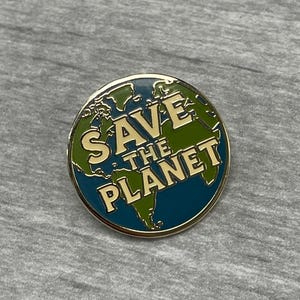 以下が含まれることがあります： 金色の縁取りと「SAVE THE PLANET」の文字が金色で書かれた丸いエナメルピン。青と緑の世界地図のデザインで、環境意識を促進しています。