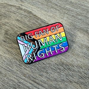 May include: A rectangular enamel pin with a rainbow flag design and the text "BIG FAN OF HUMAN RIGHTS." The pin includes the transgender pride flag and is set against a wooden background.