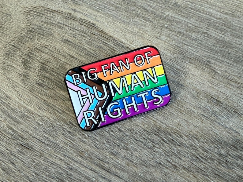 May include: A rectangular enamel pin with a rainbow flag design and the text "BIG FAN OF HUMAN RIGHTS." The pin includes the transgender pride flag and is set against a wooden background.