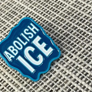 K&ouml;nnte beinhalten: Ein blauer Emaille-Pin mit den Worten "ABOLISH ICE" in wei&szlig;en Blockbuchstaben. Der Pin hat einen wellenf&ouml;rmigen, unregelm&auml;&szlig;igen Rand und ist vor einem strukturierten, grauen Hintergrund platziert.