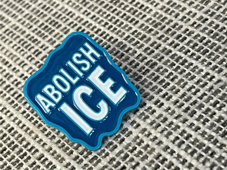 K&ouml;nnte beinhalten: Ein blauer Emaille-Pin mit den Worten "ABOLISH ICE" in wei&szlig;en Blockbuchstaben. Der Pin hat einen wellenf&ouml;rmigen, unregelm&auml;&szlig;igen Rand und ist vor einem strukturierten, grauen Hintergrund platziert.