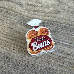 May include: A metal enamel pin shaped like a plastic bag containing three golden-brown hamburger buns. The design features a dark red label across the center with the white text That's Buns printed in a bold, stylized font against the bread graphic.