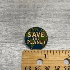 以下が含まれることがあります： 金色の縁取りが施された丸いエナメルピン。緑と青の世界地図が描かれています。「SAVE THE PLANET」の文字は白いブロック体です。ピンの直径は約2.5cmです。