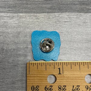 K&ouml;nnte beinhalten: Ein quadratischer, t&uuml;rkisfarbener Emaille-Anstecker mit silbernem Metallverschluss. Die Anstecknadel ist etwa 2,5 cm breit und hat einen wellenf&ouml;rmigen Rand. Die Anstecknadel liegt auf einer grauen Oberfl&auml;che neben einem Holzlineal.