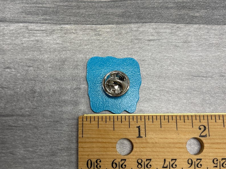 K&ouml;nnte beinhalten: Ein quadratischer, t&uuml;rkisfarbener Emaille-Anstecker mit silbernem Metallverschluss. Die Anstecknadel ist etwa 2,5 cm breit und hat einen wellenf&ouml;rmigen Rand. Die Anstecknadel liegt auf einer grauen Oberfl&auml;che neben einem Holzlineal.