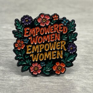 May include: A black enamel pin with the words "EMPOWERED WOMEN EMPOWER WOMEN" in red and gold lettering. The pin is surrounded by a wreath of green leaves and colorful flowers in shades of red, orange, blue, and purple.