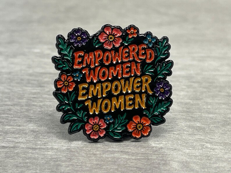 May include: A black enamel pin with the words "EMPOWERED WOMEN EMPOWER WOMEN" in red and gold lettering. The pin is surrounded by a wreath of green leaves and colorful flowers in shades of red, orange, blue, and purple.