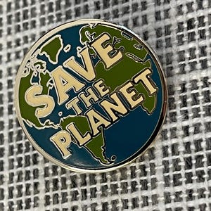 以下が含まれることがあります： 銀色の縁取りが付いた丸いエナメルピン。ピンには、緑と青の世界地図と、クリーム色の文字で「SAVE THE PLANET」という文字が描かれています。環境意識を高めるためのバッジです。