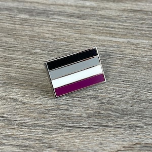 May include: A rectangular pin featuring the asexual pride flag. The flag design includes horizontal stripes in black, gray, white, and purple. The pin has a silver border and is set against a wooden background.