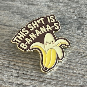 May include: A gold-colored enamel pin with the text "THIS SH*T IS B-A-N-A-N-A-S" in white letters on a brown background. The pin features a smiling cartoon banana with yellow and cream colors.