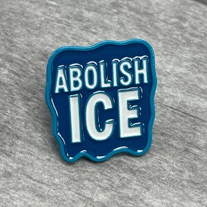K&ouml;nnte beinhalten: Blauer Emaille-Pin mit den Worten "ABOLISH ICE" in wei&szlig;en Blockbuchstaben. Der Pin hat einen wellenf&ouml;rmigen, unregelm&auml;&szlig;igen Rand und liegt vor grauem Hintergrund. Der Pin ist ein Statement-St&uuml;ck.