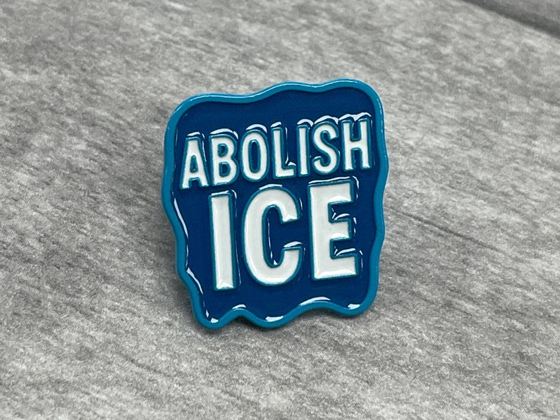K&ouml;nnte beinhalten: Blauer Emaille-Pin mit den Worten "ABOLISH ICE" in wei&szlig;en Blockbuchstaben. Der Pin hat einen wellenf&ouml;rmigen, unregelm&auml;&szlig;igen Rand und liegt vor grauem Hintergrund. Der Pin ist ein Statement-St&uuml;ck.