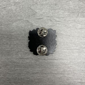 May include: A black enamel pin with two silver pin backs. The pin has a unique, irregular shape. The pin is set against a gray, textured surface.