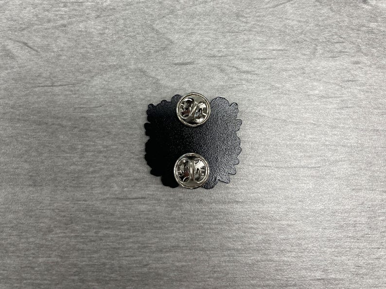 May include: A black enamel pin with two silver pin backs. The pin has a unique, irregular shape. The pin is set against a gray, textured surface.
