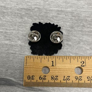 May include: A black enamel pin with two silver pin backs. The pin is approximately 1 inch in diameter and has a decorative edge. A wooden ruler is placed next to the pin for scale.