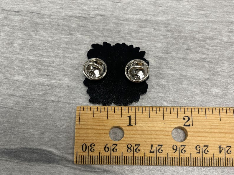 May include: A black enamel pin with two silver pin backs. The pin is approximately 1 inch in diameter and has a decorative edge. A wooden ruler is placed next to the pin for scale.