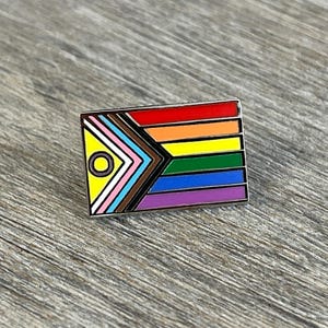 May include: A rectangular enamel pin featuring a progressive pride flag design. The pin has a rainbow stripe pattern, with additional stripes in white, pink, light blue, brown, and yellow. A black circle is on the left side.